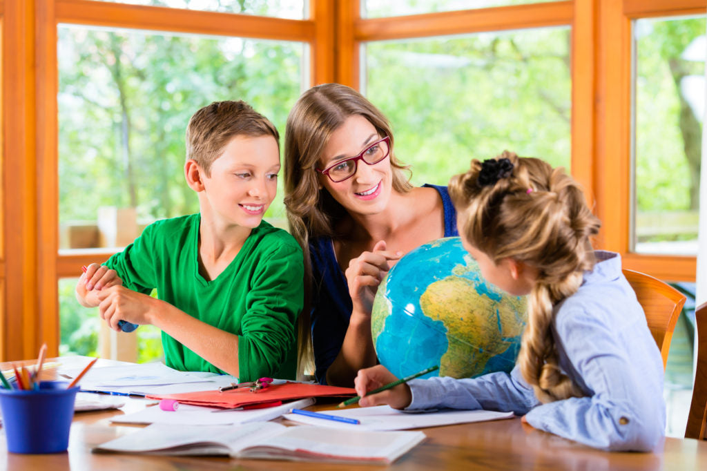 Mom pointing at a globe, homeschooling her son and daughter. Each kids has colored pencils and papers and folders on a table.